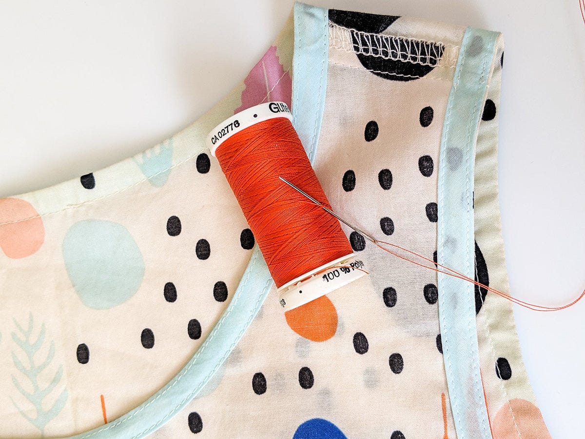 Spool of orange thread and needle resting on armhole seam of polka dot garment with blue bias binding.