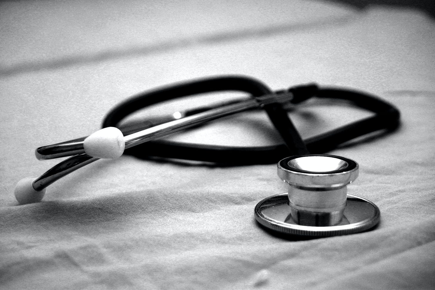 black and white photo of a stethoscope sitting on top of a bedsheet. The stethoscope is wrapped up with the metal portion in the foreground.