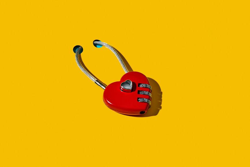 a red heart-shaped padlock locked on an orangish yellow surface with two holes a red heart-shaped padlock locked on an orangish yellow surface with two holes