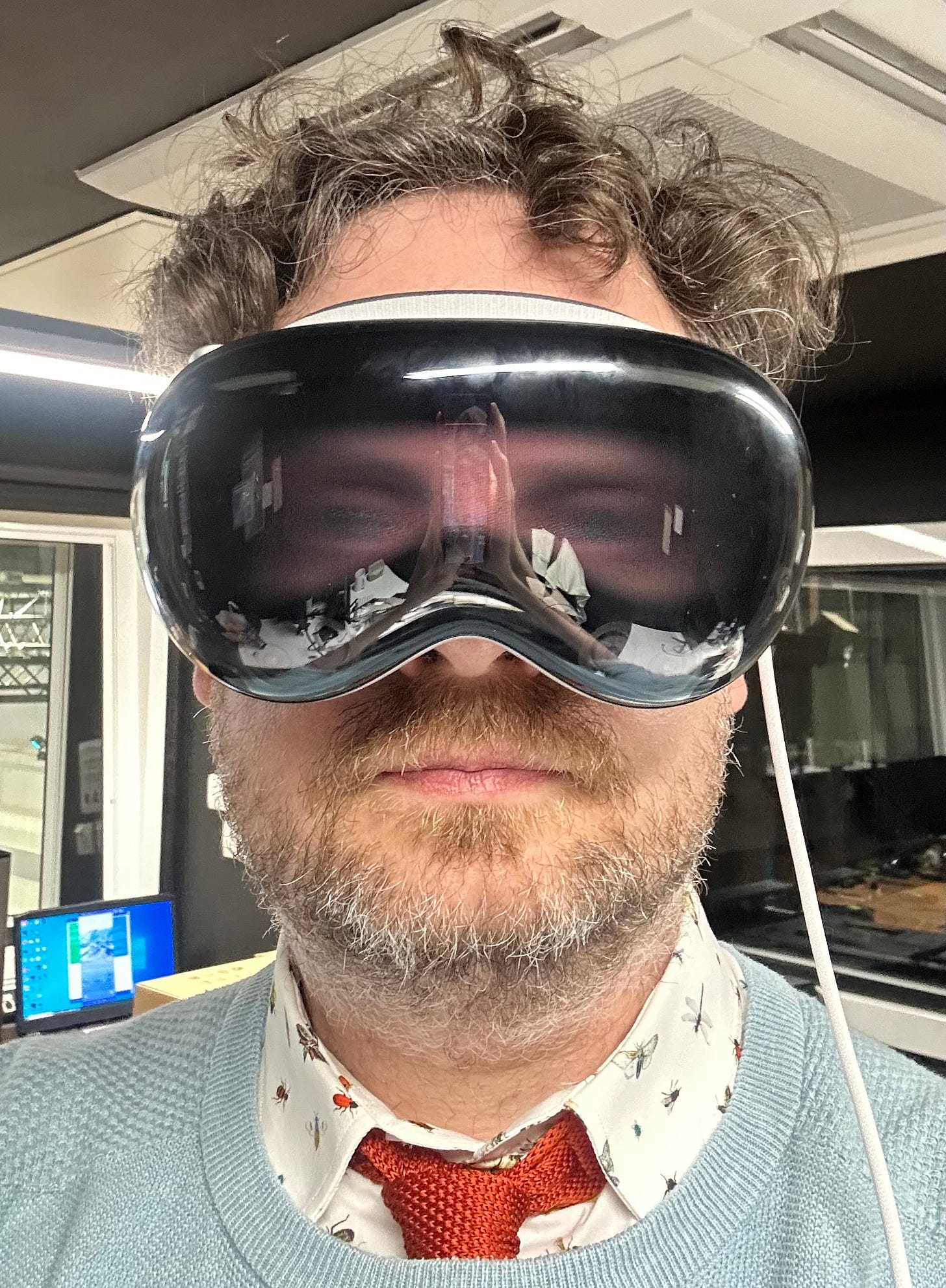 A photo of default man with curly hair and a beard wears Apple Vision Pro. He is wearing a short with insects on it under a blue jumper with an orange knitted tie. He's in an office setting.