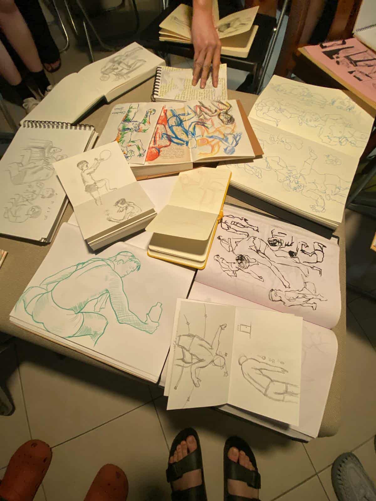 Vertical photo of many different drawings of many different styles featuring the same model, all assembled in a pile, while the feet of onlookers encircle the drawings.