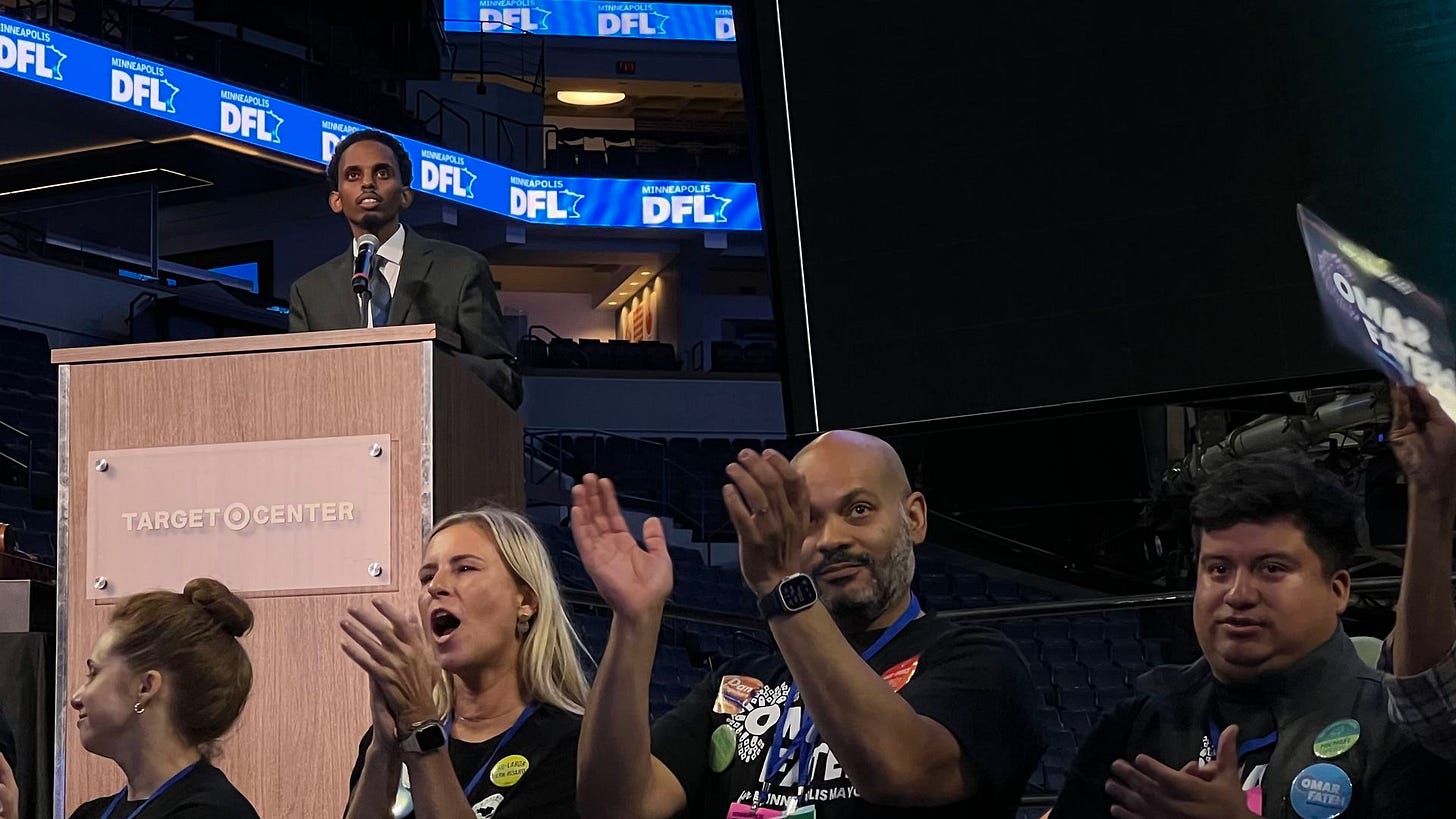 A speaker in a suit at a Target Center podium with Minneapolis DFL banners above, while four people in black shirts and campaign buttons clap and hold signs nearby.