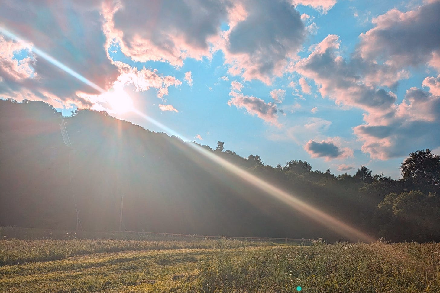 Sunbeam.jpg Sunrise over a grassy meadow with the sun cresting a tree-lined ridge. Dramatic sunbeams radiate diagonally across the frame through soft morning haze. Pink-tinged clouds scatter across a blue sky. A rustic fence runs along the field’s edge in the middle distance.