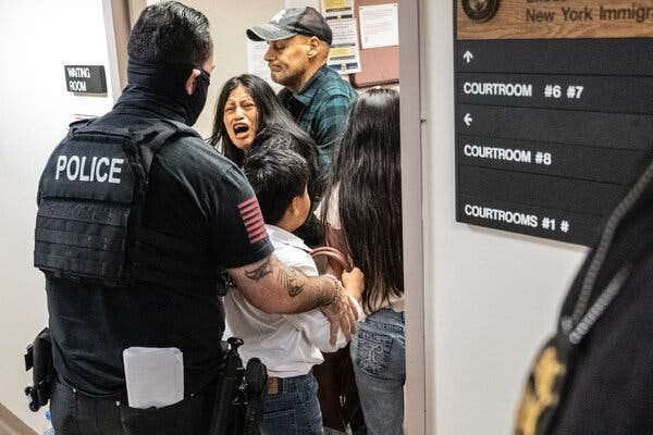 A woman is restrained by a man in a plaid shirt as a man wearing a police vest holds a little boy, and a young girl stands next to the boy.