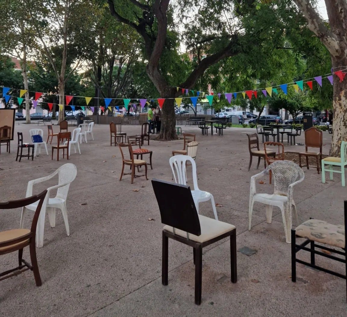 Praça Paiva Couceiro a Lisbona con numerose sedie vecchie e spaiate disposte liberamente sotto gli alberi, tra festoni colorati sospesi, creando uno spazio conviviale e informale all’aperto.