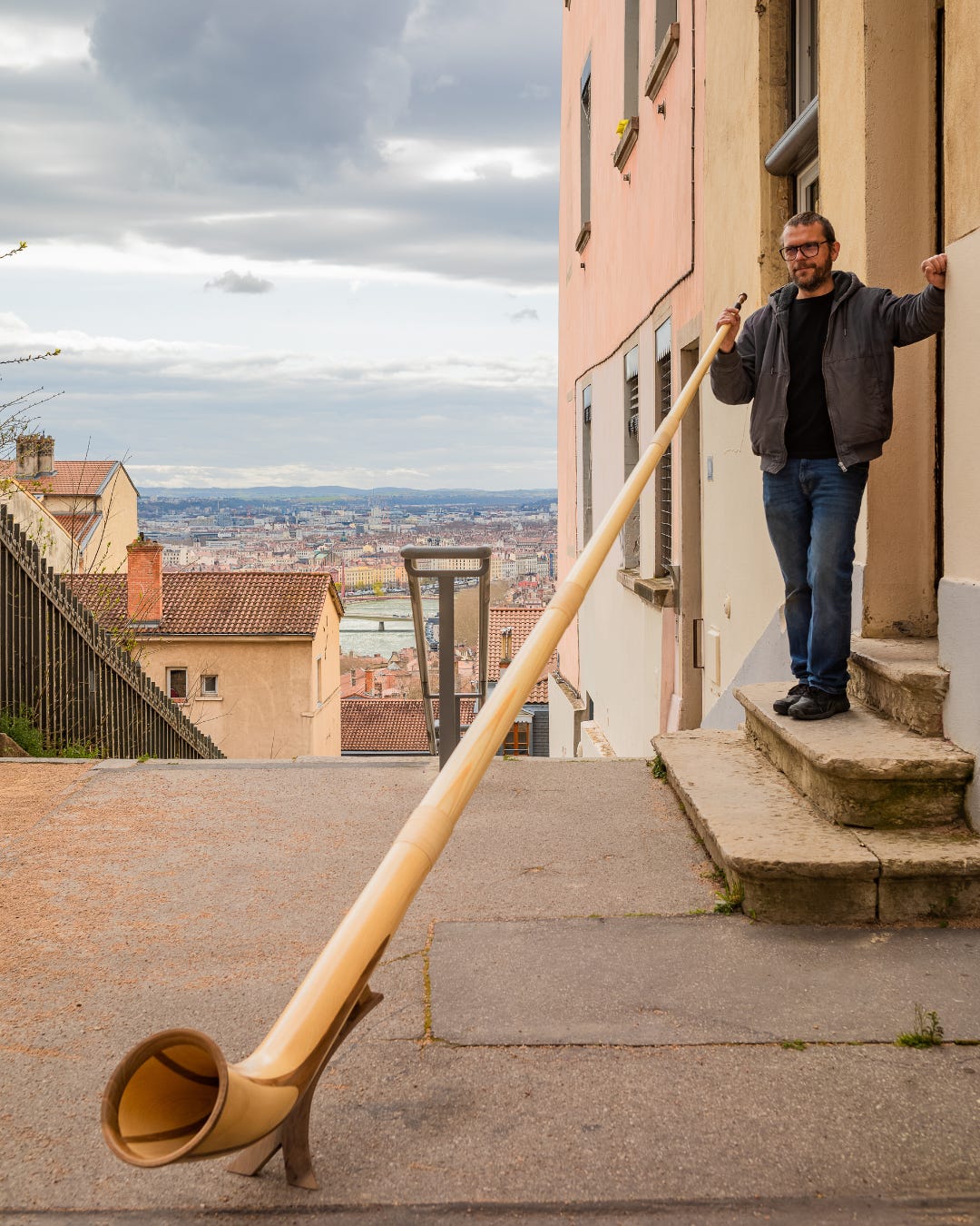 Sandro Faïta stands on steps outside a building, holding a large wooden alphorn that extends down a narrow street overlooking a cityscape.