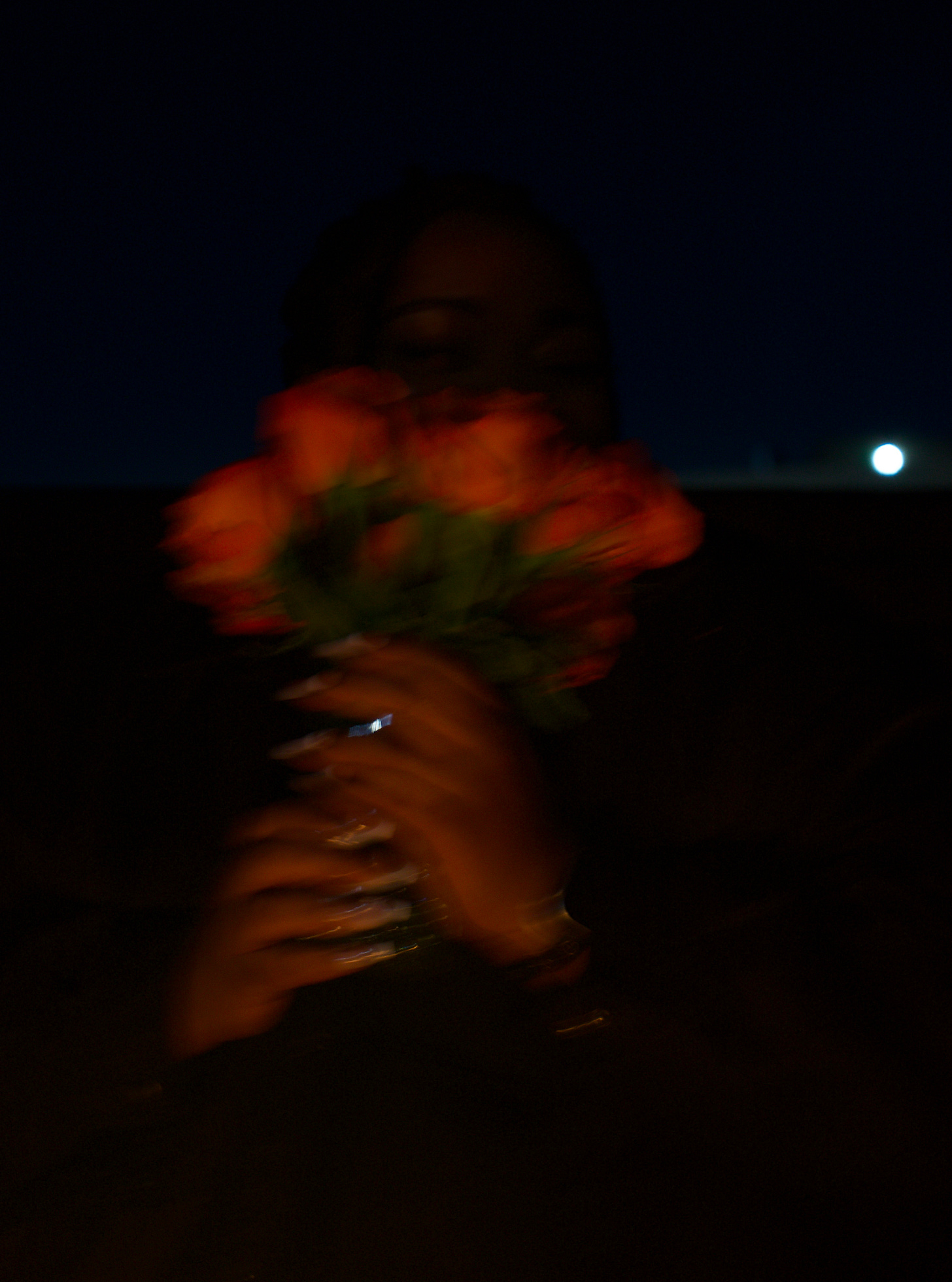 Soft-focus nighttime image of orange roses lifted toward the camera, motion blur and shallow light rendering the subject partially obscured against a dark blue sky, with subtle highlights on hands and jewelry creating a warm, atmospheric contrast.