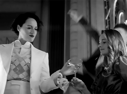 A gif of Phoebe Waller-Bridge in the Treat People With Kindness music video. She gets given a martini which she takes and starts walking in time with two dancers, her head thrown back. 