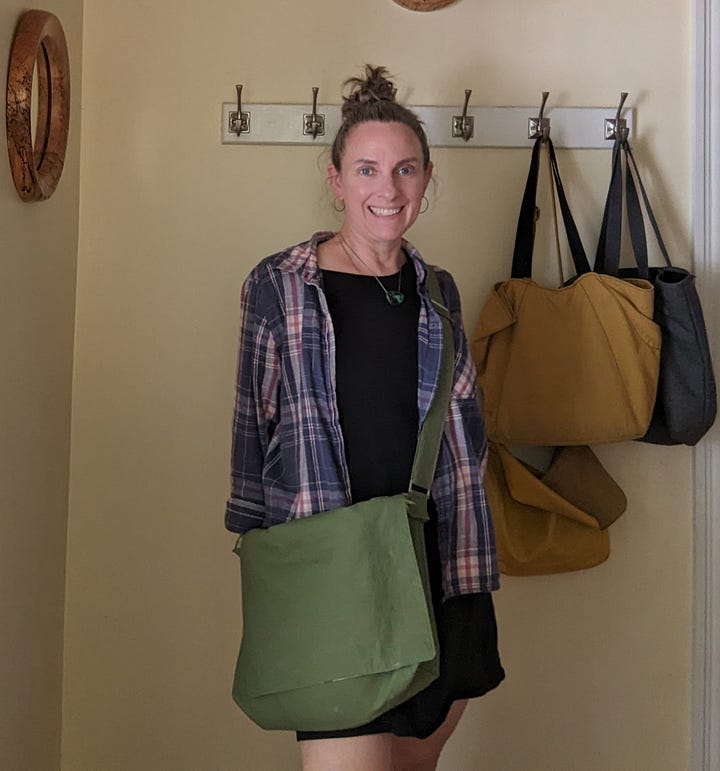 Five photos of the Mythical Messenger Bag. In the first, I (a white lady, aged 52) am standing in the hallway wearing the bag. The other photos are of the bag on its own. The bag is olive green canvas with an adjustable strap. There is hot pink decorative stitching. There is an exterior welt pocket on the back with a hot pink zipper. There is a pocket on the front with a blue square patch that says "Make Art" in a red cursive font. The inner lining fabric is floral -- blue, green, coral line drawn flowers on a white background. Inside the bag, there are two pockets. One is a zippered pocket with a hot pink zipper. The other is a divided pocket with slots for pens, a phone, and something else roughly cell-phone-sized.