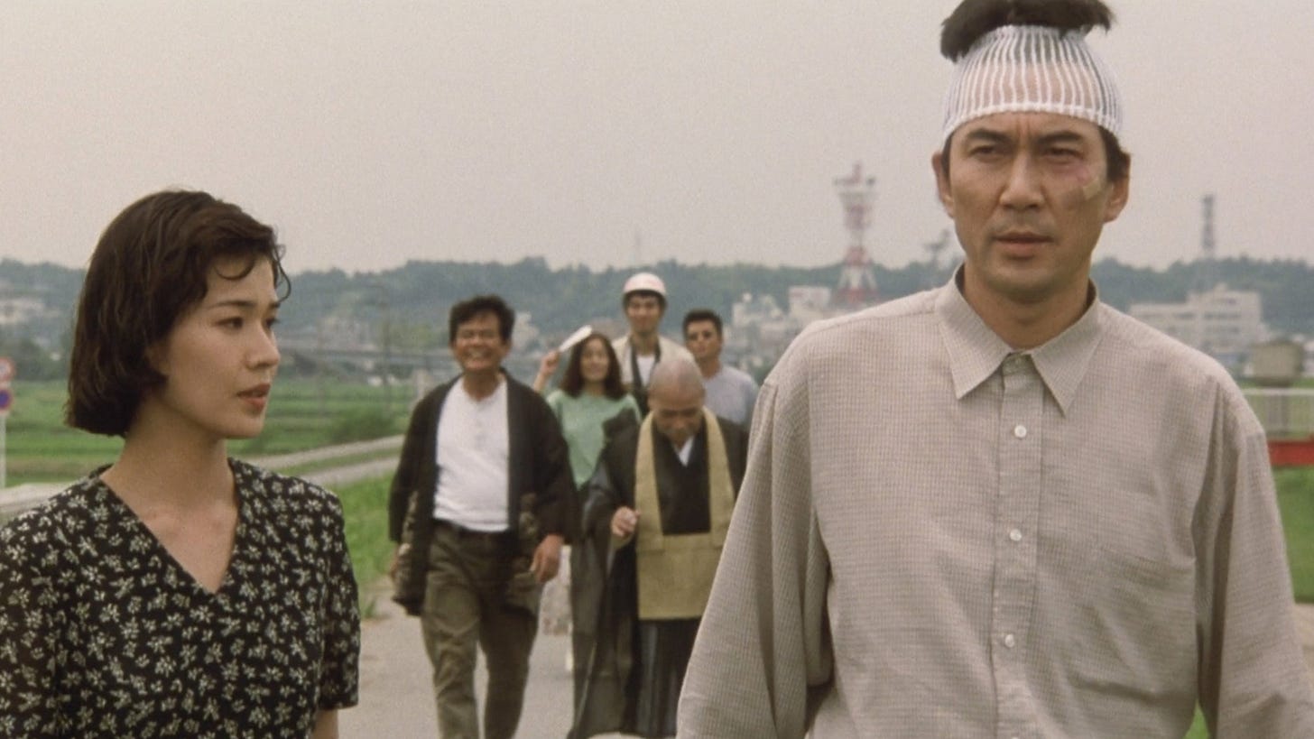 The Eel | Still features Koji Yakusho as Takuro Yamashita walking down a road with other people behind him.