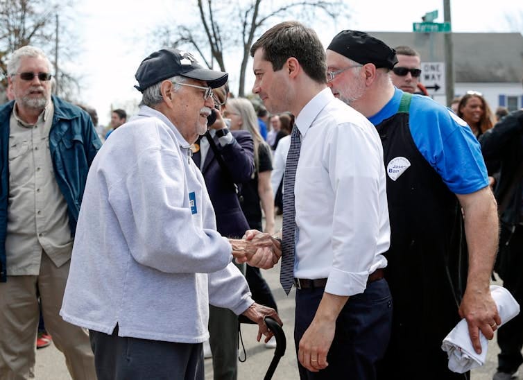 Younger man in white dress shirt and tie shakes hand with older man using cane as crowd mills around behind them