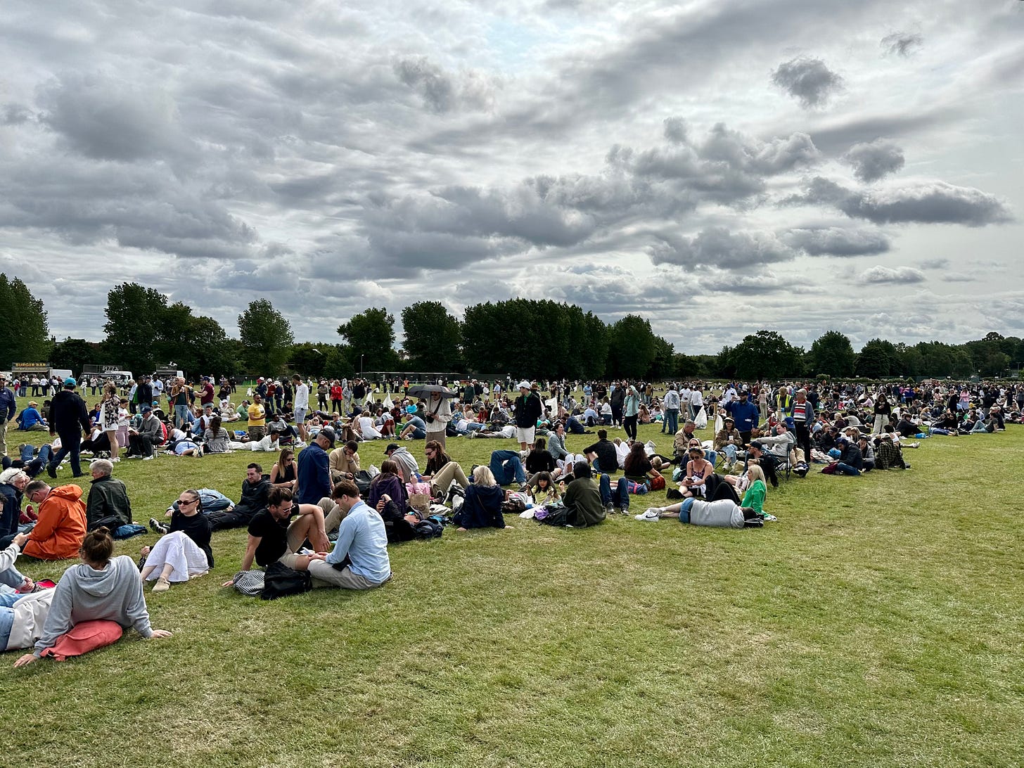 Hundreds of people sitting on the grass in Wimbledon Park waiting in The Queue under overcast skies on a July morning