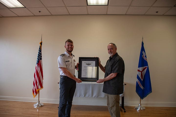 Two men stand in a plain ceremony room with wood floors, holding a framed certificate together. The man on the left wears a white Civil Air Patrol–style uniform; the man on the right wears a dark short-sleeve shirt and khaki pants. The certificate reads “Amelia Earhart Award.” An American flag is on the left and a blue Civil Air Patrol flag (with the red propeller emblem and “PATROL,” plus a “Tallahassee” streamer) is on the right. A small table with a white cloth sits behind them under fluorescent ceiling lights. Also, A man in a dark suit stands speaking in a bright room with hardwood floors and large windows overlooking an airfield. Behind him are a U.S. flag and a blue Civil Air Patrol flag. To the right is a table draped in a cloth with a framed certificate and a vase of flowers. An Epson ceiling-mounted projector hangs above, and a small bench sits by the windows.