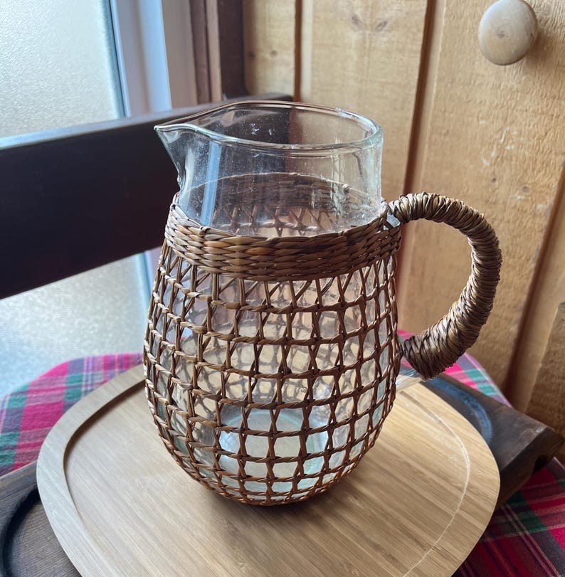 May include: A clear glass pitcher with a woven wicker handle and base. The pitcher is sitting on a light brown wooden tray.