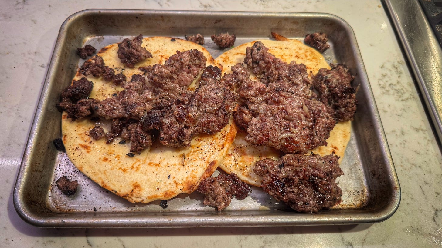 A quarter sheet pan with two pieces of naan topped with the salvaged koobideh meat. It is in browned chunks and strewn over the naan. It does not in any way resemble the skewers before they started cooking. 