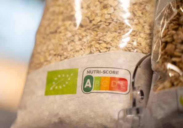 A close-up photograph titled "ffd46abc-8b04-4e6c-b627-477ff0470af5_594x415.webp" showing a transparent bag of grain or oats on a store shelf. The front-of-pack labeling features a green organic certification stamp alongside a "NUTRI-SCORE" logo, which displays a tiered color-coded nutritional scale from A to E, with a prominent dark green "A" rating highlighted. The image visually represents front-of-pack nutrition labeling policies designed to help consumers identify healthier food choices. A close-up photograph titled "ffd46abc-8b04-4e6c-b627-477ff0470af5_594x415.webp" showing a transparent bag of grain or oats on a store shelf. The front-of-pack labeling features a green organic certification stamp alongside a "NUTRI-SCORE" logo, which displays a tiered color-coded nutritional scale from A to E, with a prominent dark green "A" rating highlighted. The image visually represents front-of-pack nutrition labeling policies designed to help consumers identify healthier food choices.