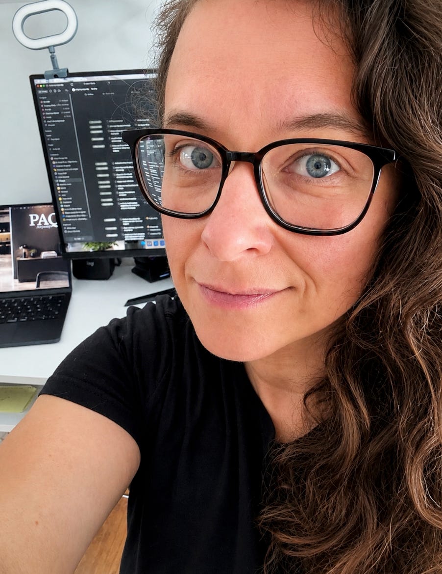 Close up professional selfie of a woman with long curly brown hair and black glasses sitting at a bright home office desk. She is wearing a black top and looking directly at the camera with a calm, confident expression. Behind her are a laptop displaying the PAQ Anyway website, a large monitor with a dark themed workspace, and a ring light. The image is sharp, well lit, and conveys a modern entrepreneur and consultant working from her office.