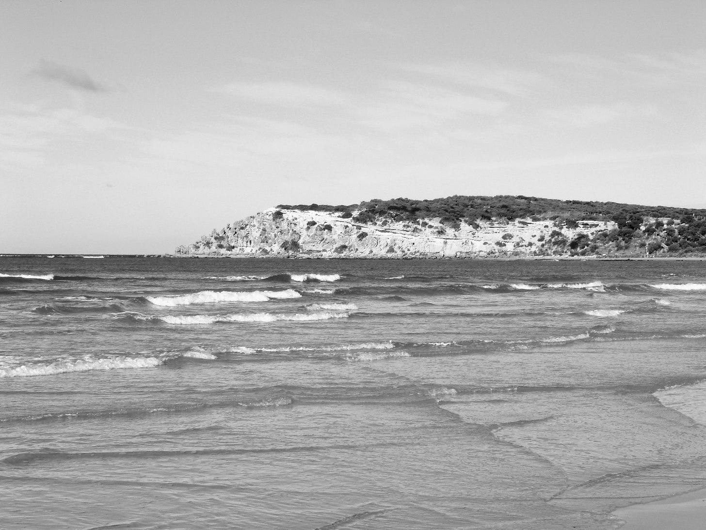 Rocky headland viewed from across a river estuary; waves breaking on the beach.