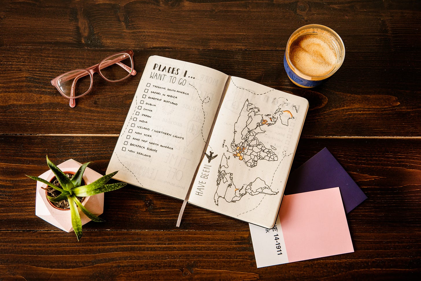 A hardwood table has a number of items: An open journal with Places I want to go, a list of places with checkboxes, and a map of the world next to the words “have been” written in it. A small houseplant, glasses, a coffee cup, and a few other assorted colored sheets of paper. A hardwood table has a number of items: An open journal with Places I want to go, a list of places with checkboxes, and a map of the world next to the words “have been” written in it. A small houseplant, glasses, a coffee cup, and a few other assorted colored sheets of paper.