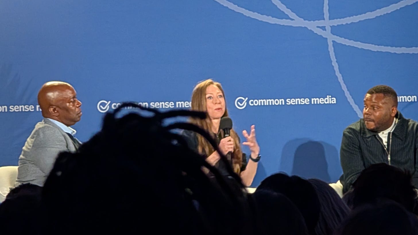 Chelsea Clinton speaks into a microphone on stage between two male panelists Chelsea Clinton speaks into a microphone on stage between two male panelists