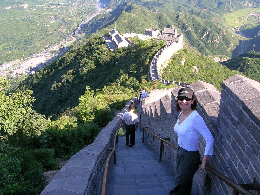A woman in a white shirt smiles while walking down a section of the Great Wall of China. The wall stretches across lush green hills under a clear sky.