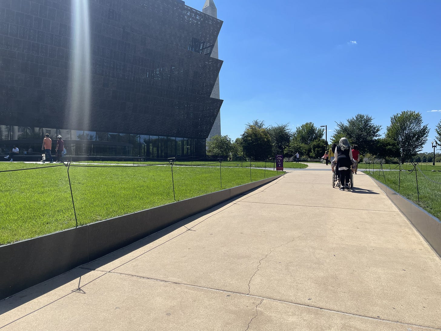 Diana pushing the author in a manual wheelchair up the wide, paved concrete ramp toward the National Museum of African American History and Culture in Washington, D.C., under bright sunlight.