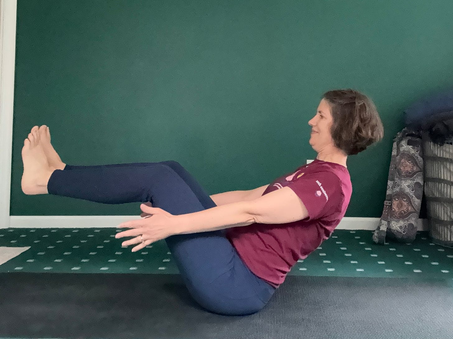 A woman sits on a black mat, leaning her torso back, lifting arms (nearly) parallel to the mat and lifting legs, so lower legs are parallel to the mat.