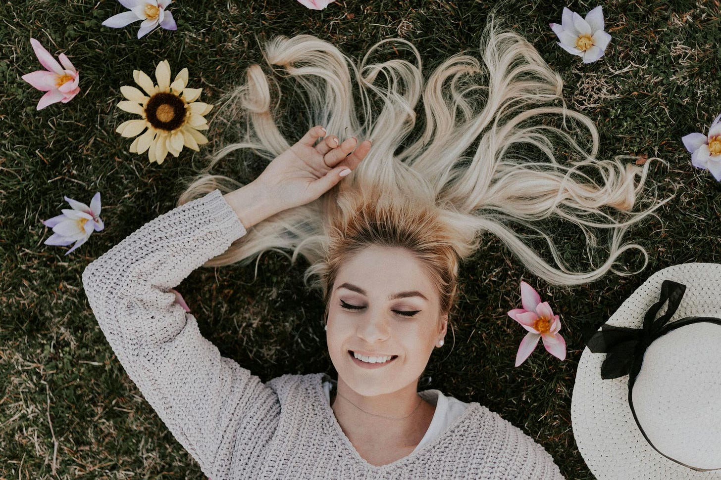 A woman lies in the grass with her eyes closed while smiling, surrounded by flower blossoms and her white hat.