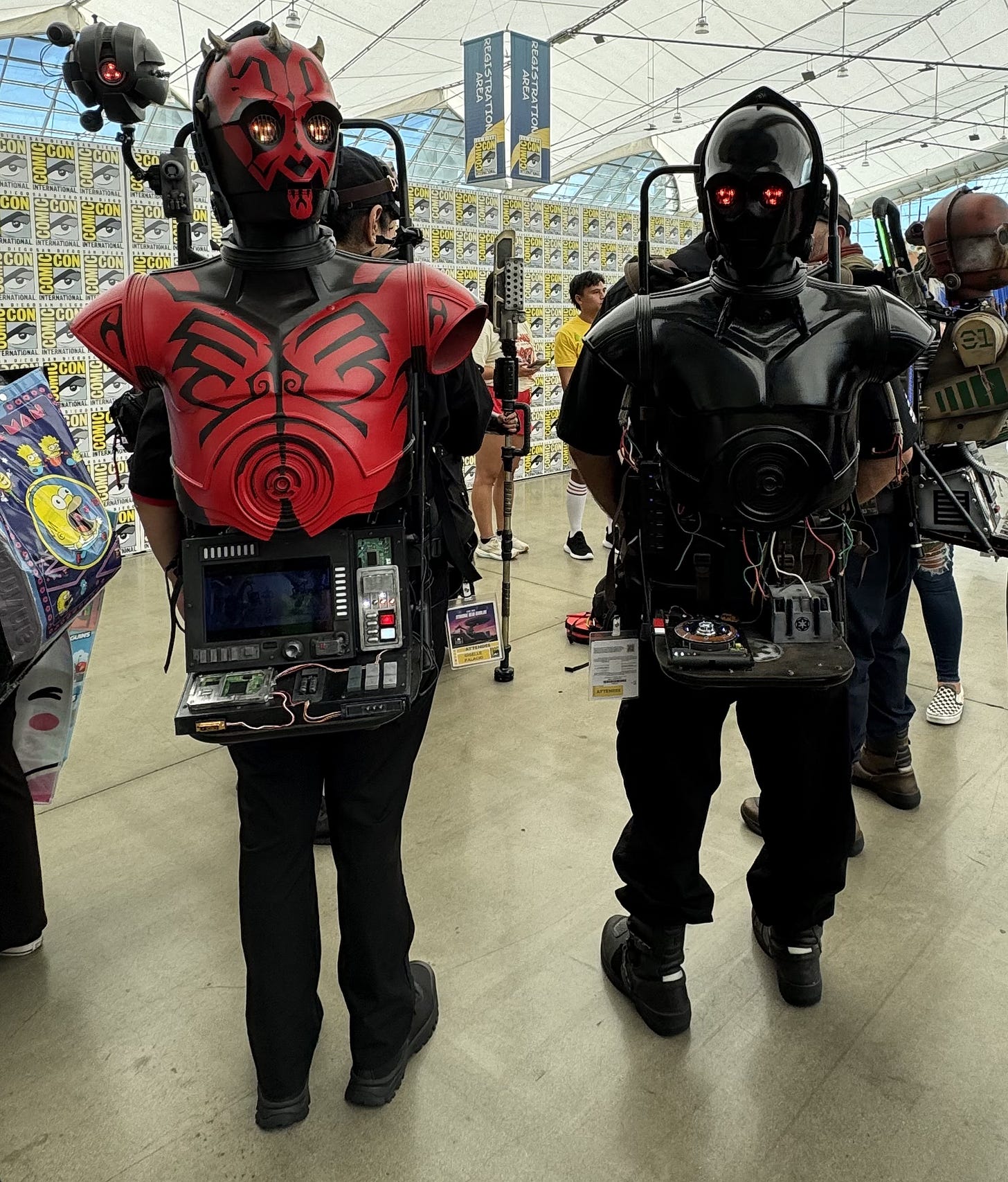 Two Comic-Con attendees wear elaborate droid-inspired costumes featuring illuminated red eyes, sculpted armor, exposed electronics, and detailed control panels built into their torsos. One costume is finished in red and black with horn-like details, while the other is glossy black. A small hovering droid prop floats beside them, and other costumed attendees fill the bustling registration area in the background.