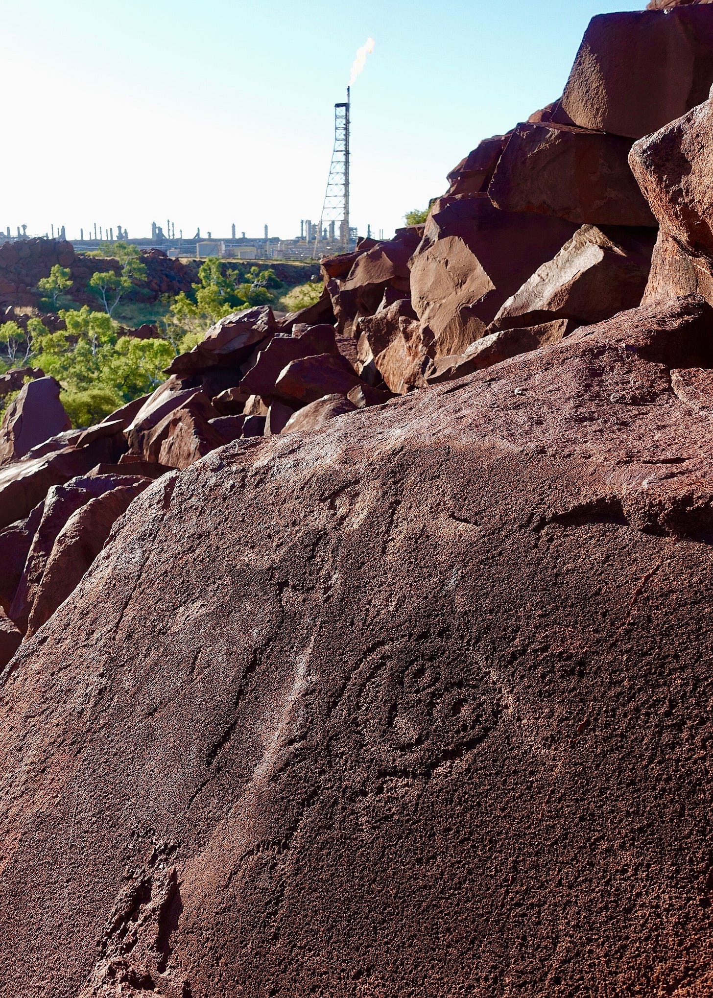 A dark red boulder carved with a pecked spiral motif in the foreground. Behind a jumble of red rocks and green scrub, a tall industrial flare tower rises against a pale sky. A dark red boulder carved with a pecked spiral motif in the foreground. Behind a jumble of red rocks and green scrub, a tall industrial flare tower rises against a pale sky.