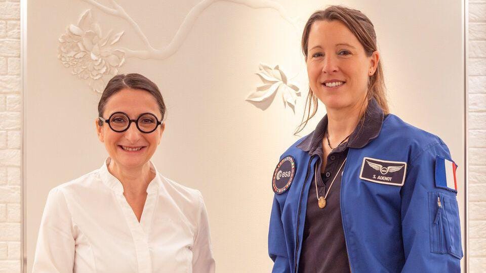 Anne-Sophie Pic, wearing a white shirt and her signature black round glasses, and Sophie Adenot, wearing a blue Nasa jacket, stand together smiling. Anne-Sophie Pic, wearing a white shirt and her signature black round glasses, and Sophie Adenot, wearing a blue Nasa jacket, stand together smiling.