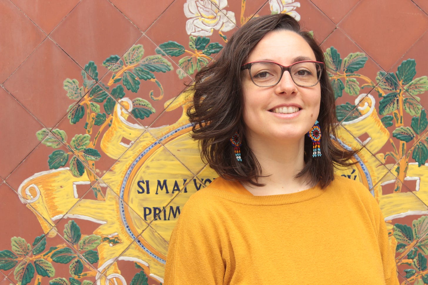 Alice Orrù sorride di fronte alla camera: indossa degli occhiali grandi con montatura rossa e nera, degli orecchini multicolore di artigianato messicano e un maglione color ocra. Dietro di lei, una parete color terracotta decorata con motivi floreali gialli e verdi.