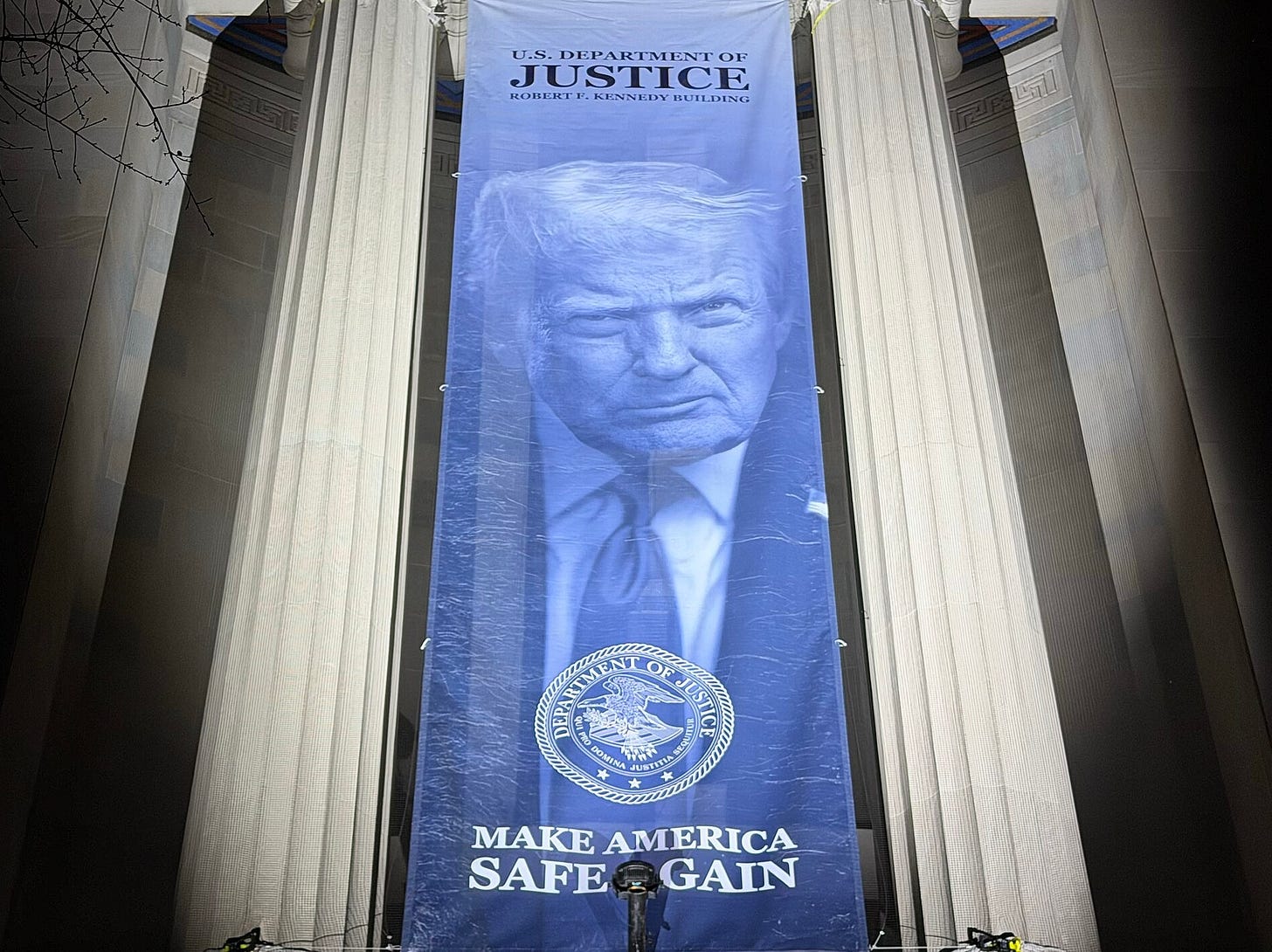 Large blue banner with Donald Trump's image and the seal of the Department of Justic reading, "U.S. Department of Justice Robert F. Kennedy Building Make America Safe Again" hanging between Ionic columns. Large blue banner with Donald Trump's image and the seal of the Department of Justic reading, "U.S. Department of Justice Robert F. Kennedy Building Make America Safe Again" hanging between Ionic columns.