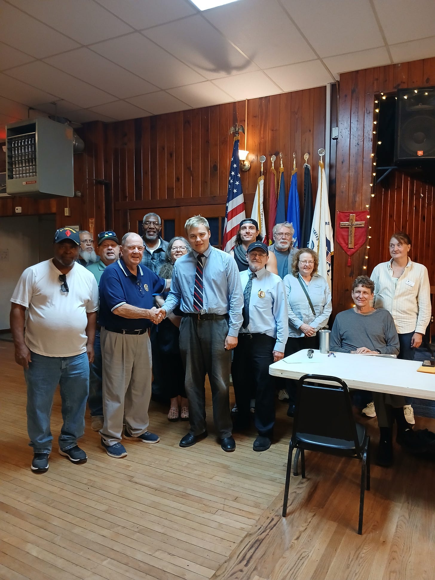 A group of about fifteen people poses in a wood‑paneled community hall with a hardwood floor. At the center, an older man in a navy polo and cap shakes hands with a younger man in a light blue dress shirt and red striped tie. Behind them are the U.S. flag and several military service flags on stands. To the right is a white folding table with a seated person, a tumbler, and papers, plus an empty chair. String lights, a wall speaker, and overhead ceiling tiles are visible, creating a casual meeting or recognition event atmosphere. A group of about fifteen people poses in a wood‑paneled community hall with a hardwood floor. At the center, an older man in a navy polo and cap shakes hands with a younger man in a light blue dress shirt and red striped tie. Behind them are the U.S. flag and several military service flags on stands. To the right is a white folding table with a seated person, a tumbler, and papers, plus an empty chair. String lights, a wall speaker, and overhead ceiling tiles are visible, creating a casual meeting or recognition event atmosphere.