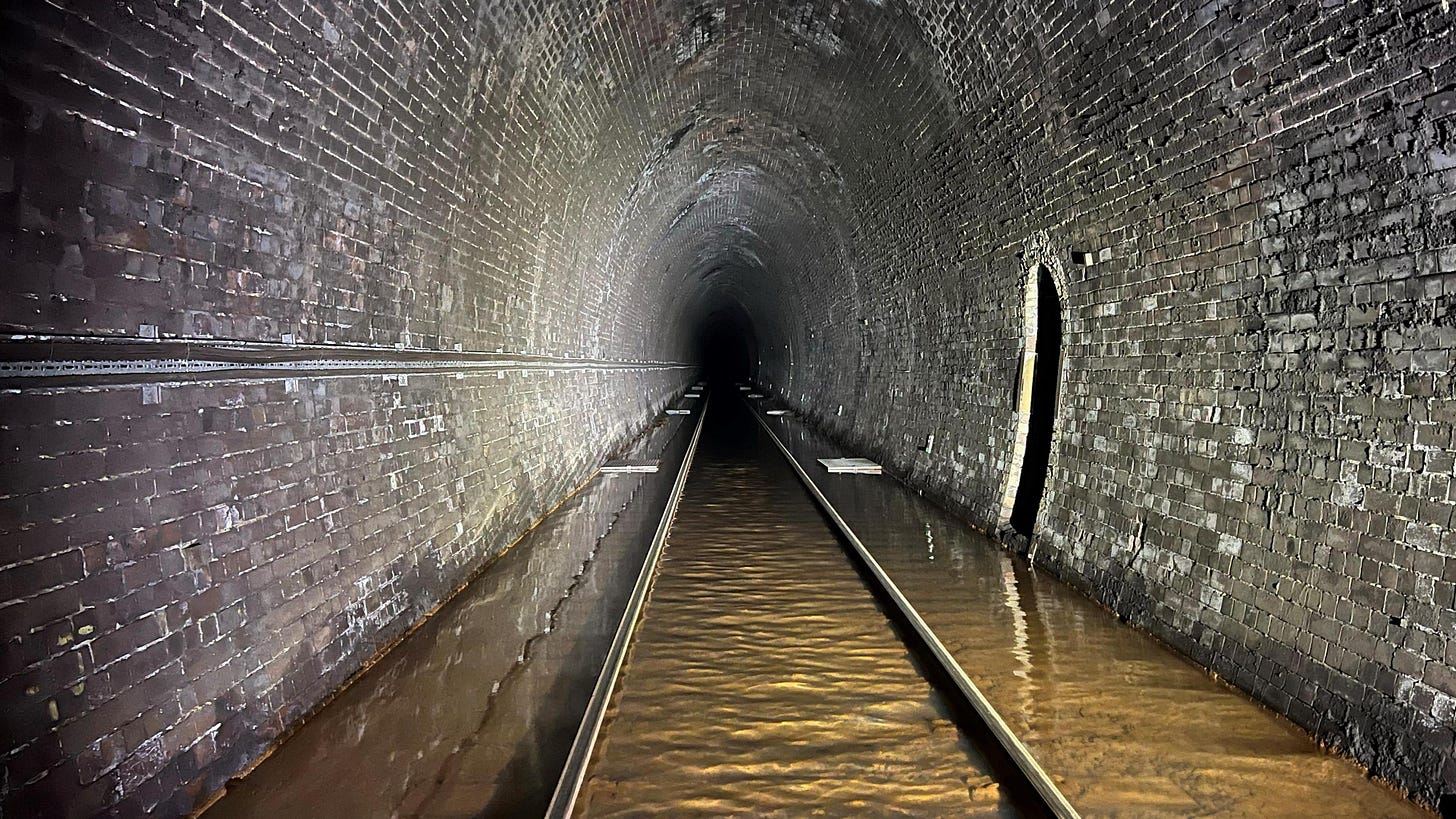 Flood water in Whitehaven tunnel containing iron ochre. The tunnel has a single rail track running along it, which is poking above the layer of water. The curved walls are made of grey brick. Flood water in Whitehaven tunnel containing iron ochre. The tunnel has a single rail track running along it, which is poking above the layer of water. The curved walls are made of grey brick.