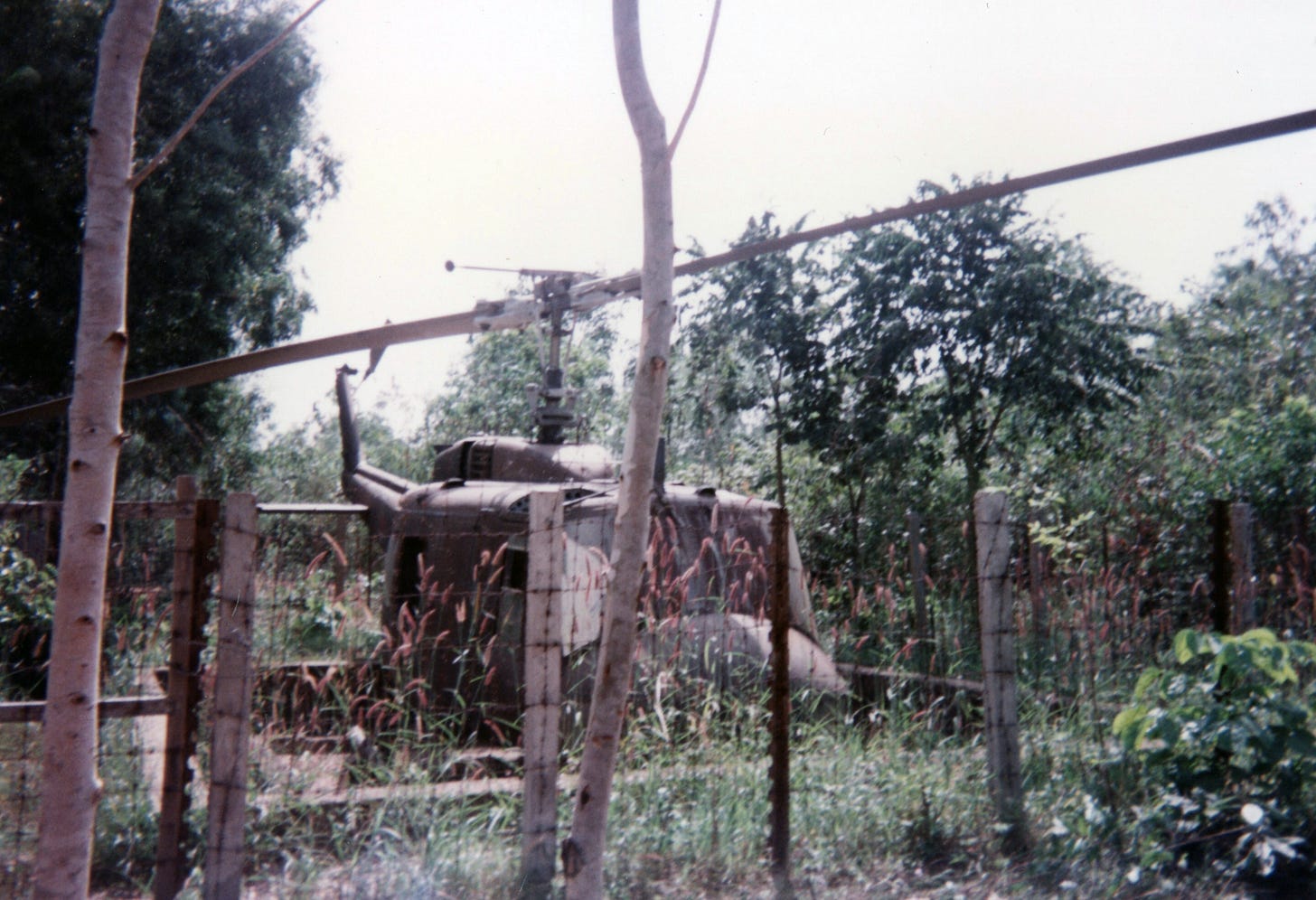 Army helicopter burned out and sitting behind a barbed-wire fence in the jungle. The photo is old and the colors faded to muted greys, greens, and browns.