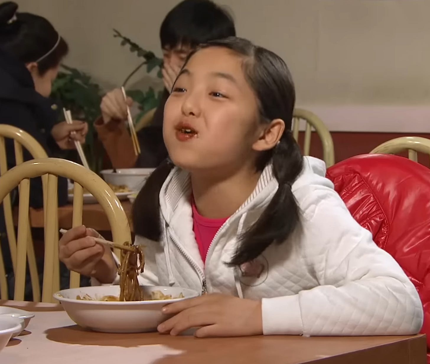 A young girl in a white jacket sits at a restaurant table eating jjajangmyeon, Korean-Chinese black bean noodles, with chopsticks. Other people are seated behind her, and a red jacket hangs on the chair beside her.