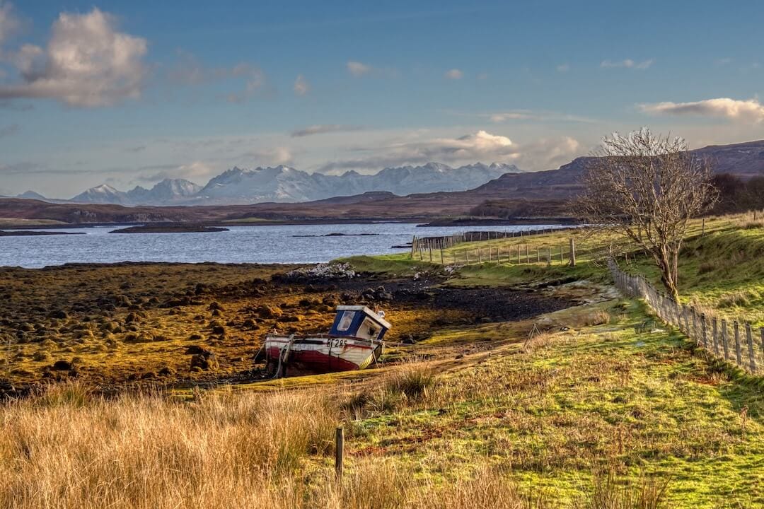 A small fishing boat in a field with a loch and snow-capped mountain range in the background
