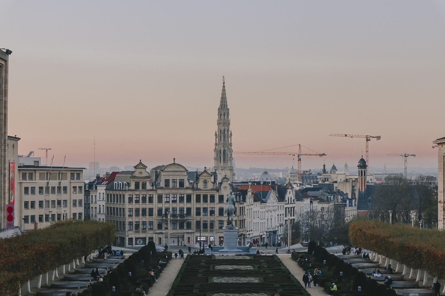 Mont des Arts in Brussels at sunset, with a pink-hued sky framing the spire of the Brussels Town Hall and terraced gardens below.
