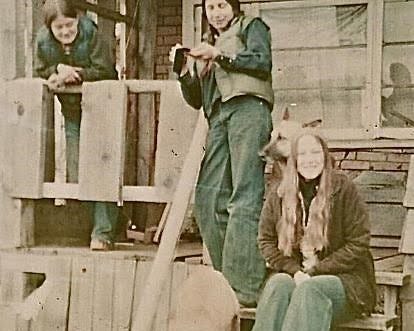 An old photo of three people sitting, standing and slouching in front of a wooden home. An old photo of three people sitting, standing and slouching in front of a wooden home.