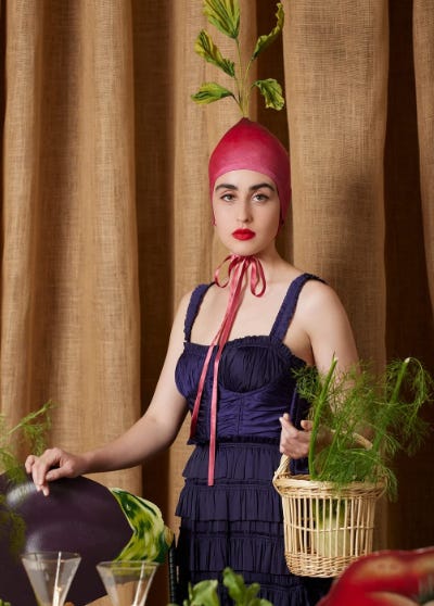 light skinned woman wearing a radish hat and holding vegetables in a basket posing by a table of vegetables