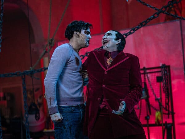 Nicolas Cage, as a bloody, grinning Dracula in a red velvet suit, has one arm around Nicholas Hoult, who wears a bloody blue sweatshirt and jeans and clenches his fist with an angry expression. They stand in front of chains, ropes and an illuminated red background.