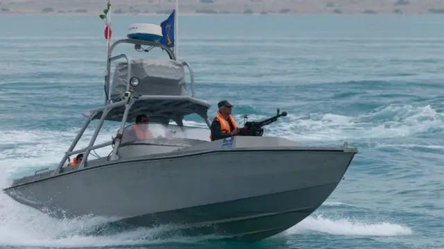 A boat patrols the waters off an arid coast, with a man in a bright orange jacket onboard holding a machine gun