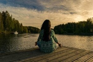 Woman on a dock, facing a body of water, sitting in the lotus position presumably meditating