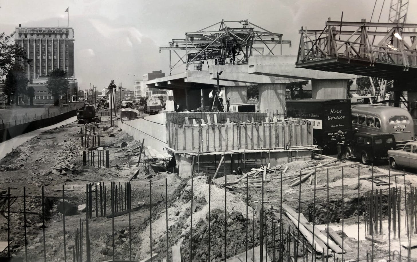 Black and white photograph of a chaotic construction site. A viaduct is being built on the right with formwork for concrete to be poured; in the centre a ramp is half-built. A traffic jam snakes through the worksite. Black and white photograph of a chaotic construction site. A viaduct is being built on the right with formwork for concrete to be poured; in the centre a ramp is half-built. A traffic jam snakes through the worksite.
