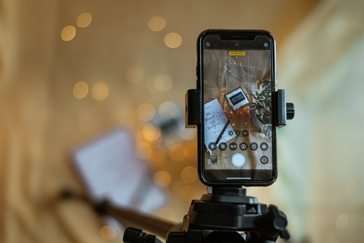 A smartphone held upright in a tripod mount with its camera app open, in front of a softly blurred, warmly lit background.