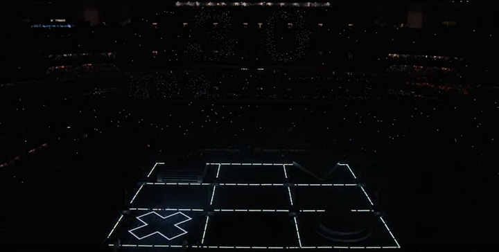 First image: An overhead view of 28 cardboard boxes filled with crouching Black men dressed in all black. Kendrick Lamar's cornrowed hair stands out at the center of these boxes. Second image: Nine large square stages are lit in a much larger, dark stadium.