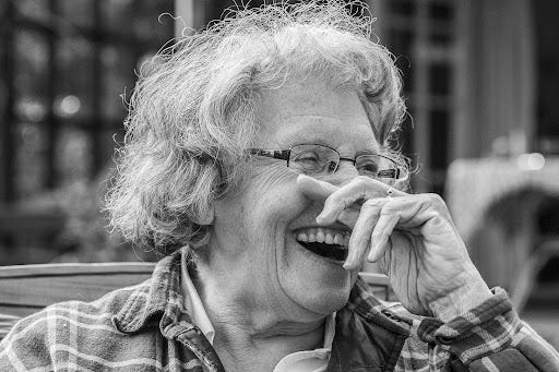 Black and white portrait photo of older white woman with gray hair and glasses as she attempts to cover up a hearty laugh with her left hand. Black and white portrait photo of older white woman with gray hair and glasses as she attempts to cover up a hearty laugh with her left hand.