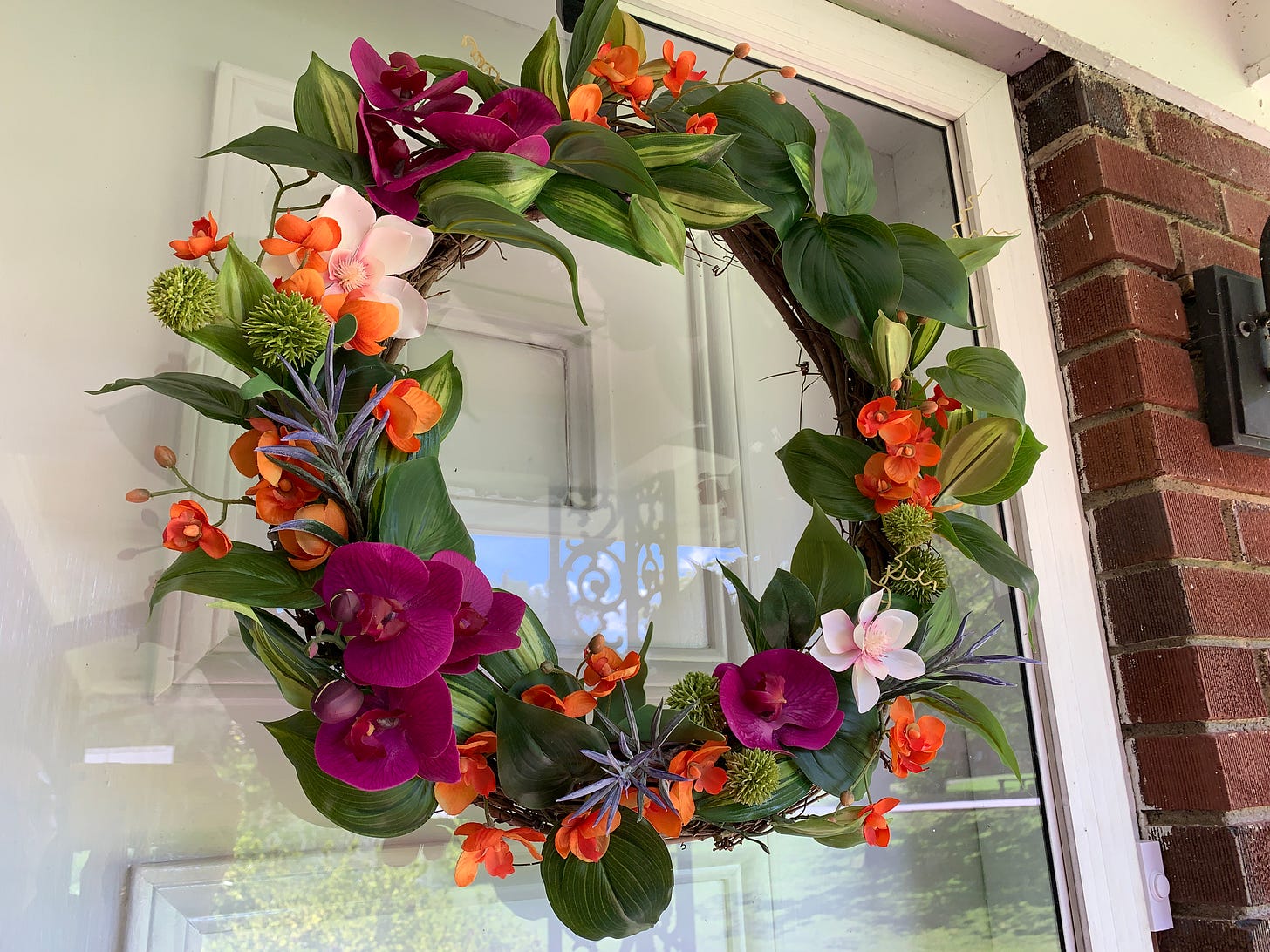 Hawaiian-inspired handmade wreath hanging from a glass storm door. Flowers include purple orchids, leafy greens found on a lei, and other pink, orange, and green flowers the creator doesn't know the names of.