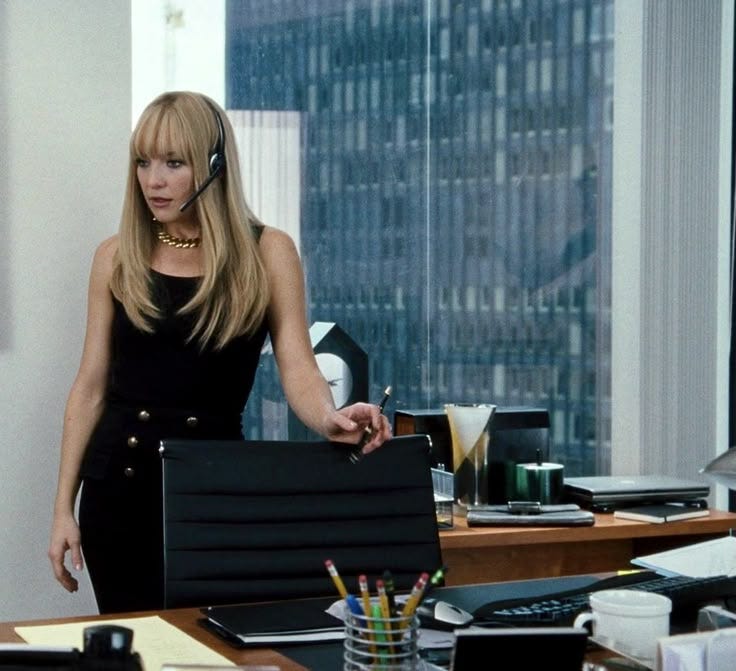 This may contain: a woman standing in an office holding a black box and talking on the phone with another person behind her